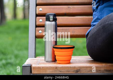 Termo de acero y taza de silicona. Platos turísticos en el banco. Tomar café en el parque. Foto de stock