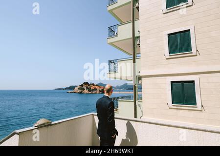 Hombre elegante con traje negro se encuentra en un balcón con vistas a la isla Sveti Stefan cerca de Budva, Montenegro. Vista posterior Foto de stock