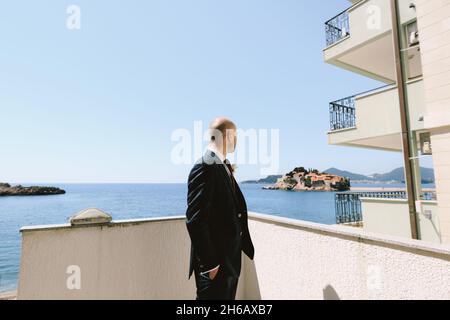 Elegante novio en un traje negro con un boutonniere se encuentra en el balcón con vistas a la isla de Sveti Stefan cerca de Budva, Montenegro Foto de stock