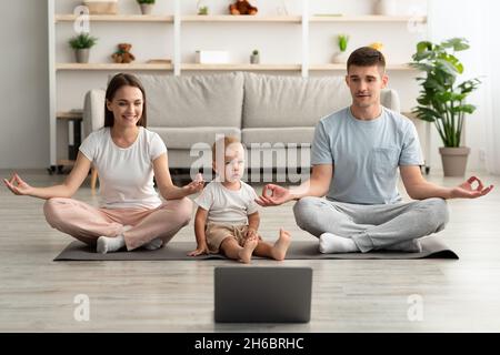 Yoga en línea. Feliz Familia Joven con Niño Infantil Meditando Juntos En Casa Con Computadora Portátil, Padres Cheerful Sentados En Posición De Loto, Viendo Video Foto de stock