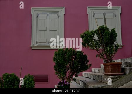 Fachada de edificio neoclásico con una pared de estuco rosa goma burbuja, persianas de madera gris y plantas de albahaca en los escalones de piedra de Nafplio, Grecia. Foto de stock