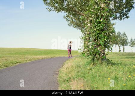Longitud completa de jogger hembra adulto activo con cola larga en ropa deportiva que corre a lo largo de camino de asfalto en la zona rural en día soleado en Islandia Foto de stock