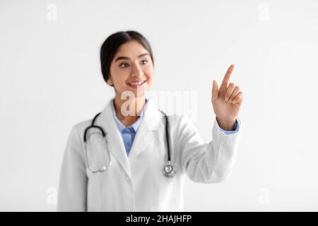 Sonriente joven hindú doctor en abrigo blanco con stetoscopio apunta el dedo al espacio en blanco Foto de stock