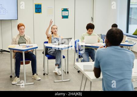 Un grupo diverso de niños sentados en los escritorios en el salón de clase de la escuela moderna se centra en la niña adolescente levantando la mano, el espacio de copia