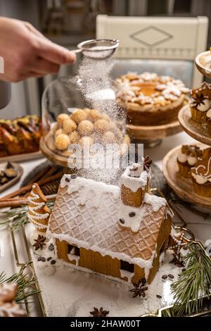 Mano de mujer rociando azúcar glaseado sobre casa de pan de jengibre. Mesa navideña festiva con dulces Foto de stock