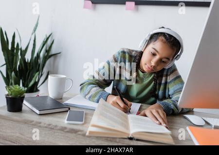 una niña afroamericana con auriculares escribiendo cerca del libro y la computadora mientras estudiaba en casa