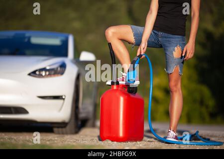 Chica de pie al lado de un coche eléctrico. Sujetando el cable de carga y el contenedor de gascolina. Foto de stock