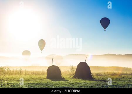 Silueta de globos de aire caliente preparándose para el lanzamiento en una mañana foggy detrás de dos aystacks. Foto tomada el 3nd de octubre de 2021 cerca de un campo en Rogoz, Ma