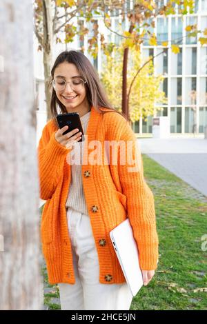 Joven positiva hembra en gafas y cárdigan naranja con smartphone mientras está de pie ordenador portátil cerca del árbol en el parque Fotografía de - Alamy