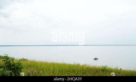 Campo verde y un lago tranquilo con bote de goma flotante. Hierba verde fresca y flores en la orilla del gran lago sobre fondo cielo nublado en el summ Foto de stock