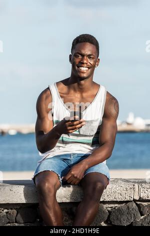 Feliz hombre negro con ropa de verano sentado en una valla de piedra contra el mar en Lanzarote y navegar por el smartphone mientras disfruta del día de verano y mira a ca Foto de stock