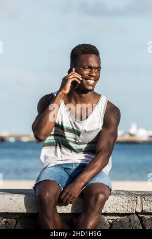 Sonriente hombre negro con traje de verano sentado en una valla de piedra junto al mar y hablando por teléfono móvil mientras disfruta de un fin de semana en Lanzarote y mirando hacia fuera Foto de stock