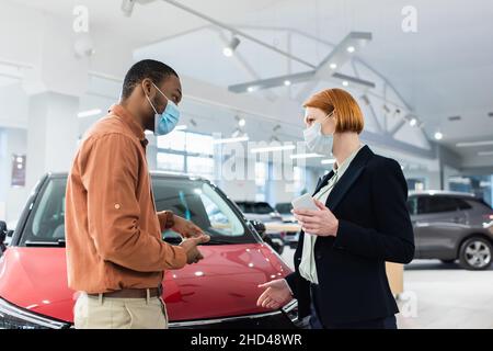 hombre afroamericano con máscara médica apuntando con las manos al coche cerca del concesionario de automóviles Foto de stock