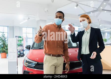 concesionario de automóviles y cliente afroamericano en máscaras médicas apuntando con los dedos en la sala de exposición de automóviles Foto de stock
