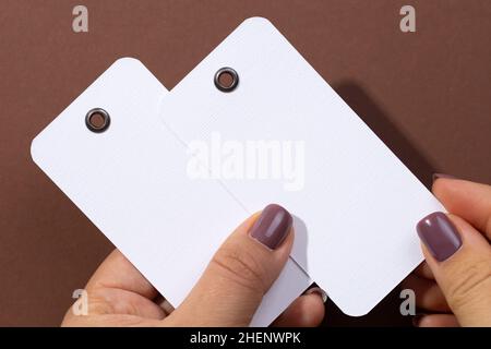 Dos etiquetas blancas en blanco de forma rectangular hechas de cartón con  pequeños agujeros en la parte superior para ropa en manos femeninas en el  centro del fondo marrón Fotografía de stock -