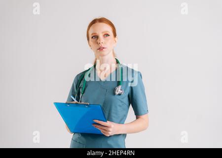 Retrato de una joven enfermera pensante en la prescripción de la escritura del uniforme médico en el portapapeles, llena la historia médica, mirando hacia arriba, Foto de stock