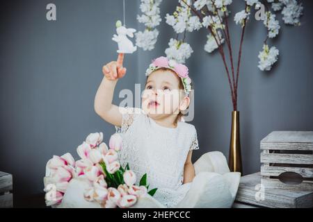 Retrato de una niña en un vestido blanco con un borde de flor en su cabeza en un huevo decorativo con tulipanes en el tema de Pascua. Flores de fondo Foto de stock