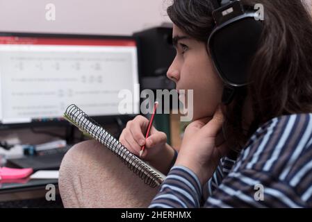 Adolescente transgénero con auriculares y micrófono estudiando delante de la computadora, mira un vídeo de su tutor para resolver tareas matemáticas, mientras se toca Foto de stock