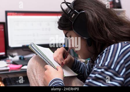 Adolescente transgénero con auriculares y micrófono estudiando delante de la computadora desenfocada, escuchando música mientras resolviendo tareas matemáticas en ella no Foto de stock
