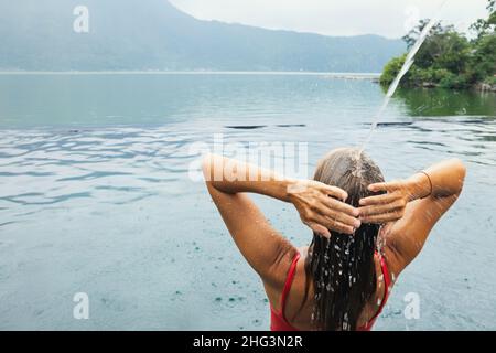 Una mujer joven se relaja bajo el agua corriente en la piscina infinita con hermosas vistas al lago. Balneario natural de aguas termales junto al volcán Batur. Tour turístico en Kintaman