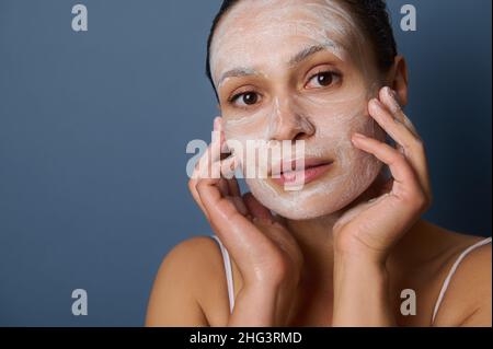 Primer plano de una joven mujer de piel oscura muy joven que se da un masaje facial, eliminando maquillaje y limpiando su cara con una limpieza facial exfoliante bea Foto de stock