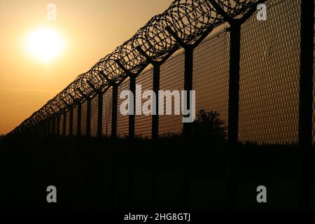 Alambre de púas en la parte superior de la valla de malla metálica que rodea el aeropuerto contra el sol de la tarde Foto de stock
