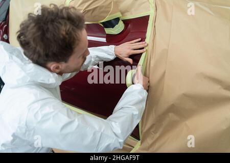 Auto pintor enmascarando la carrocería del coche con cinta y papel antes de volver a pintar. Mecánico en un traje blanco de protección trabaja en un garaje