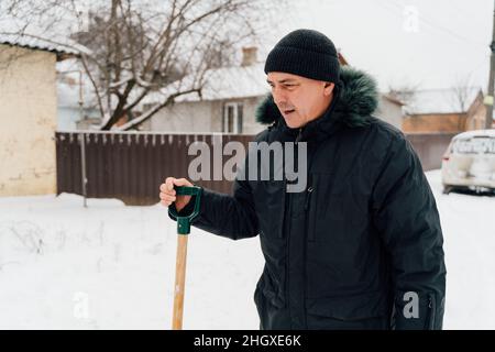 Caída de nieve. Hombre mayor limpiando nieve en invierno con una pala en un patio durante el día frío. Concepto de problemas de invierno Foto de stock