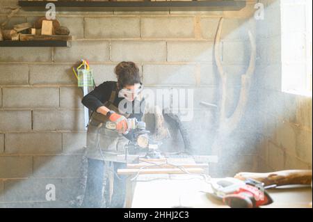 Artesana que lleva guantes, delantal y gafas haciendo un detalle de madera con una amoladora eléctrica en un banco de trabajo en un taller de artesanía