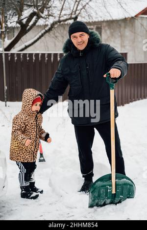 Caída de nieve. Hombre mayor con niño limpiando nieve en invierno con una pala en un patio durante el día frío. Concepto de problemas de invierno Foto de stock