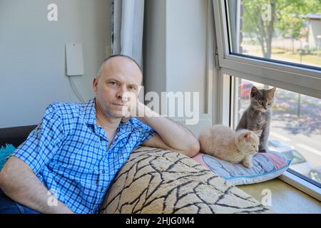 Retrato de un hombre maduro y dos gatitos esponjosos en un sofá junto a la soleada ventana Foto de stock