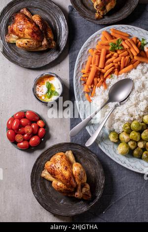 Mesa de cena con pollo a la parrilla, arroz y verduras, coles de bruselas al horno, zanahoria en un plato oval grande y platos de invitados en mantel de lino azul o Foto de stock