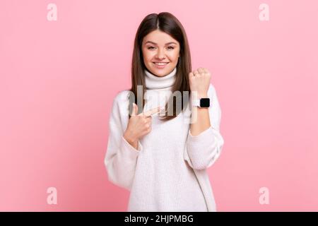 Alegre mujer morena apuntando reloj de pulsera y sonriendo, mostrando el tiempo de reunión acordado, minutos felices, usando suéter blanco de estilo informal. Estudio en interior grabado aislado sobre fondo rosa. Foto de stock