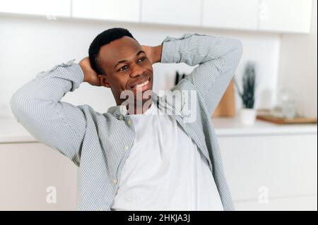 Retrato de un hermoso hombre afroamericano. Hombre negro atractivo vestido con ropa informal con estilo sentado en casa en la sala de estar en una postura relajada con las manos detrás de la cabeza, sonriendo felizmente Foto de stock