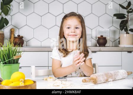 Retrato de una niña sonriente sentada en una silla, amasándose, haciendo galletas de diferentes formas. Preparación de la masa, eje de rodadura con harina. Esculpir forma b Foto de stock