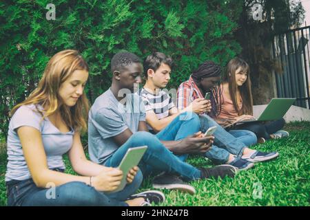 Estudiantes multiétnicos de secundaria relajándose usando aparatos electrónicos al aire libre Foto de stock
