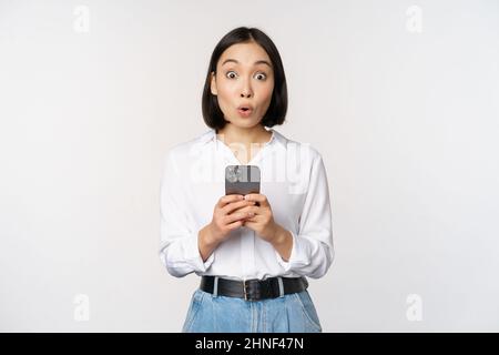 Concepto de tecnología. Retrato de mujer asiática con teléfono móvil, chica sosteniendo smartphone y reaccionando sorprendido en la notificación de teléfono móvil, información de la aplicación Foto de stock