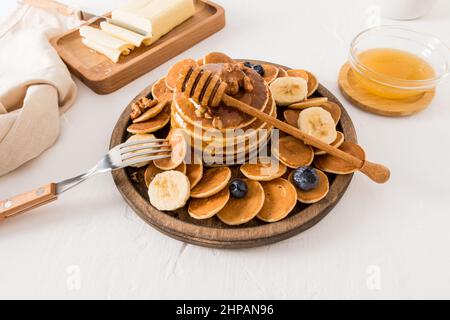 tortitas caseras en un plato redondo de madera con rodajas de plátano, nueces, bayas. miel en un tazón, mantequilla en el tablero para mejorar el sabor Foto de stock