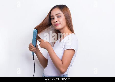 joven asiático hermosa mujer feliz alisando el pelo con plancha para el pelo sobre fondo blanco Foto de stock