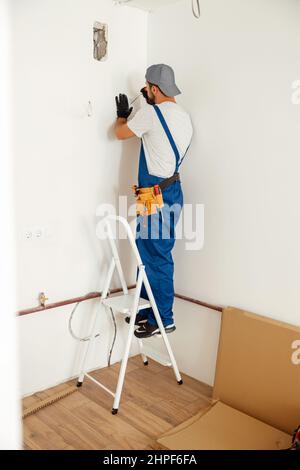 Electricista, hombre de mano parado en una escalera mientras trabaja en la instalación del enchufe en la pared de la casa Foto de stock