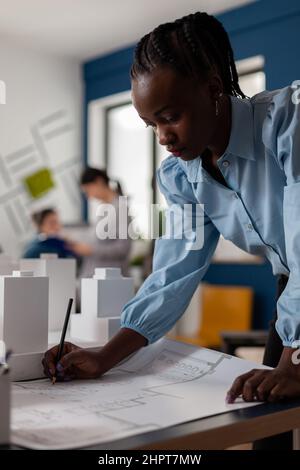 Retrato del arquitecto haciendo notas sobre los documentos de los planos de construcción en el escritorio de la moderna oficina de arquitectura. Diseño de planos de diseño de ingeniero de desarrollo para proyecto residencial junto a la maqueta de espuma.