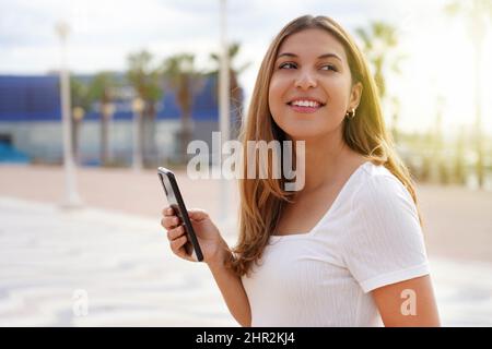 Chica sonriente caminando sola sosteniendo el teléfono al atardecer en verano Foto de stock