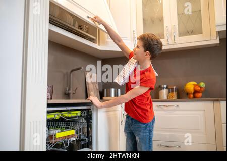 Niño contento mirando la vajilla limpia en el estante secante Foto de stock