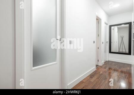 Pasillo de una casa residencial con pisos de madera marrón, paredes blancas prístinas, y un espejo enmarcado en negro Foto de stock