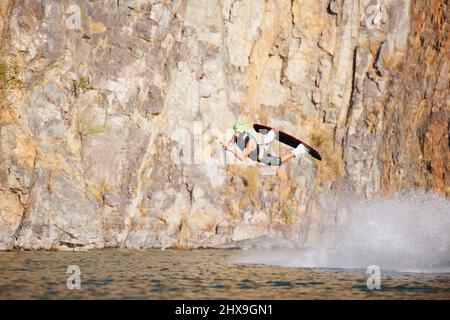 Un hombre joven con un wakeboard helmut y chaleco salvavidas en un lago. Un hombre joven con casco y chaleco salvavidas wakeboarding en un lago.