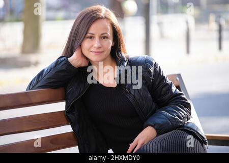 Joven mujer feliz sentada relajada en el banco de la calle en un cálido día de autarmo. Bienestar y descanso del concepto de prisa cotidiana. Foto de stock