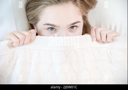 Una joven mujer atractiva apesta debajo de las mantas escondidas detrás de ellas con miedo cuando se despierta. El concepto de mujer Foto de stock