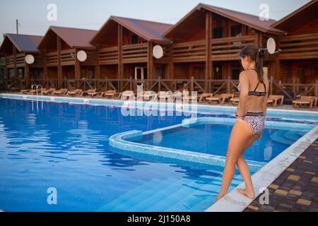 Una chica de pelo oscuro con un pelo largo y fluido en un traje de baño de leopardo brillante está probando agua fresca y limpia y transparente en la piscina, bajo un verano