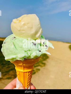 A mano sosteniendo un gran cono de helado con chip de menta y vainilla en un soleado día de cielo azul en la playa Foto de stock