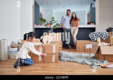 una pareja joven abrazó de pie frente al escritorio de la cocina, sonriendo, mirando a su daugter en un apartamento nuevo con cajas de cartón desempaquetadas Foto de stock